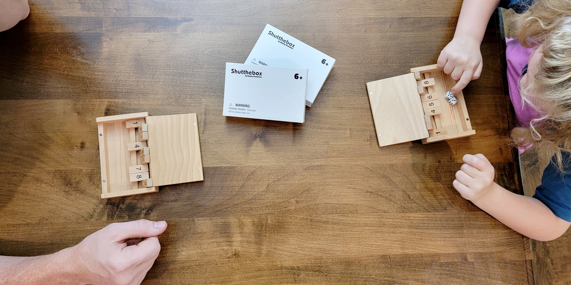 Overhead image of a parent and a child sitting at a wooden table with open Shut the Box by Velocity Promotions dice games playing a two-player game of Shut the Box